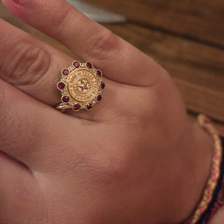 TXST Class Ring | A gold signet-style ring with red gemstone accents worn on a person's finger, shown against a warm wooden background.