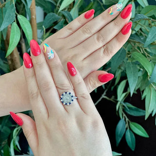 UTSA Custom Class Ring | A close-up of a person's hands with bright red manicured nails and a colorful floral nail design on one finger, wearing a silver ring with blue stones. The background is filled with green leaves.