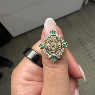 Baylor Class Ring | A close-up of a hand holding a Baylor Graduation gold ring with a central clock face motif, surrounded by green emerald-cut stones and small clear stones, with a black band on the other visible finger.