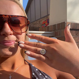 Custom College Class Rings | A close-up selfie of a woman wearing tortoiseshell sunglasses, a black-and-white polka-dot top, and a silver cross necklace, holding up her hand to display a silver ring with a round clear gemstone.