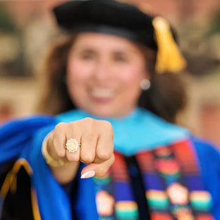 UTSA Class Ring | A close-up lifestyle shot of a woman in a blue and black academic gown and cap pointing directly at the camera, wearing a gold ring on her finger. The background is heavily blurred, showing an outdoor setting.