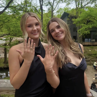 Custom College Class Rings | Two women posing outdoors in front of trees, both wearing black dresses and displaying gold graduation rings on their hands.