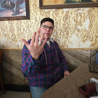 Tarleton University Men's Class Ring | A man wearing glasses and a blue and red plaid hoodie sits in a restaurant booth, holding up his hand to show off a large silver ring on his index finger. The background features a textured gold wallpaper and framed paintings.
