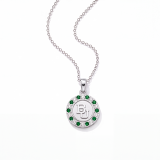 A silver pendant necklace featuring a circular medallion with a white 'BU' monogram in the center, surrounded by a halo of small green gemstones and a border of clear stones, suspended from a silver ball chain on a white background.