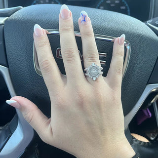 Tarleton Class ring | A close-up of a hand resting on a car steering wheel, wearing a silver-toned ring with a round sunburst design and clear stones. The hand has long, manicured nails with light pink polish and one blue accent nail.