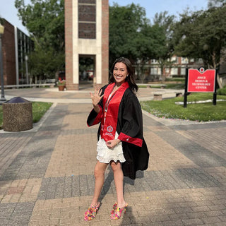 Texas Tech Class Ring | A woman wearing a black graduation gown with red trim and a matching red sash stands outdoors in front of a brick clock tower on a paved campus walkway.