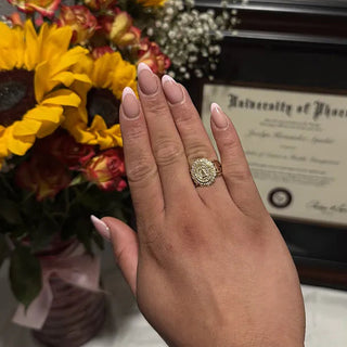 University of Houston Class Ring | A hand with manicured nails wearing a gold ring with a floral or paisley pattern, shown in a lifestyle setting with a bouquet of yellow and red flowers and a framed diploma in the background.
